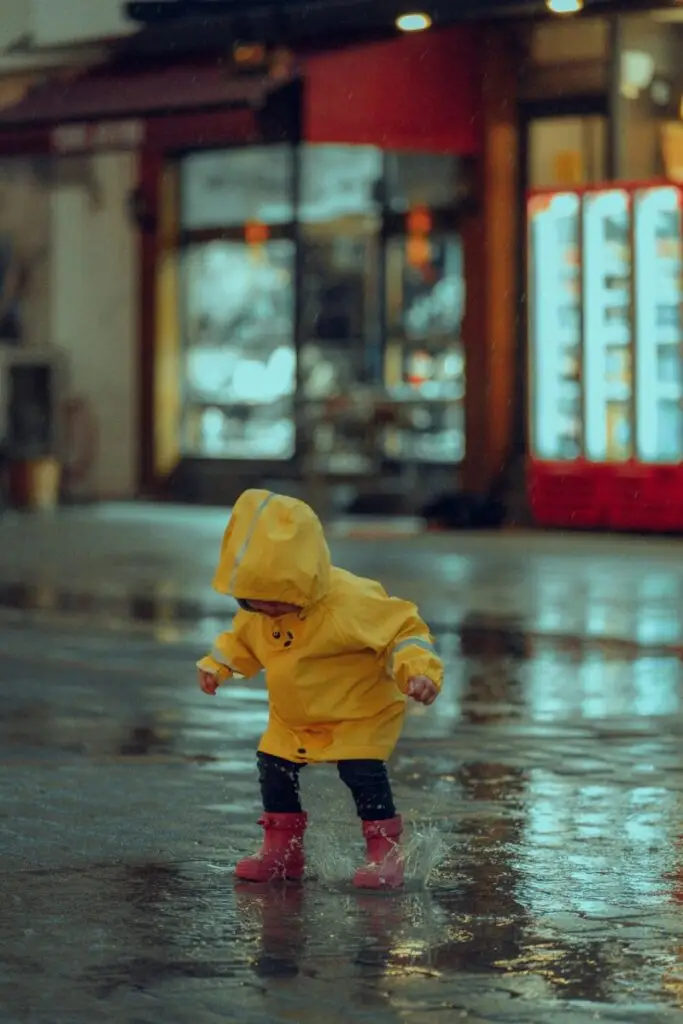 A young child in a yellow raincoat joyfully jumps in a puddle on a rainy night street.