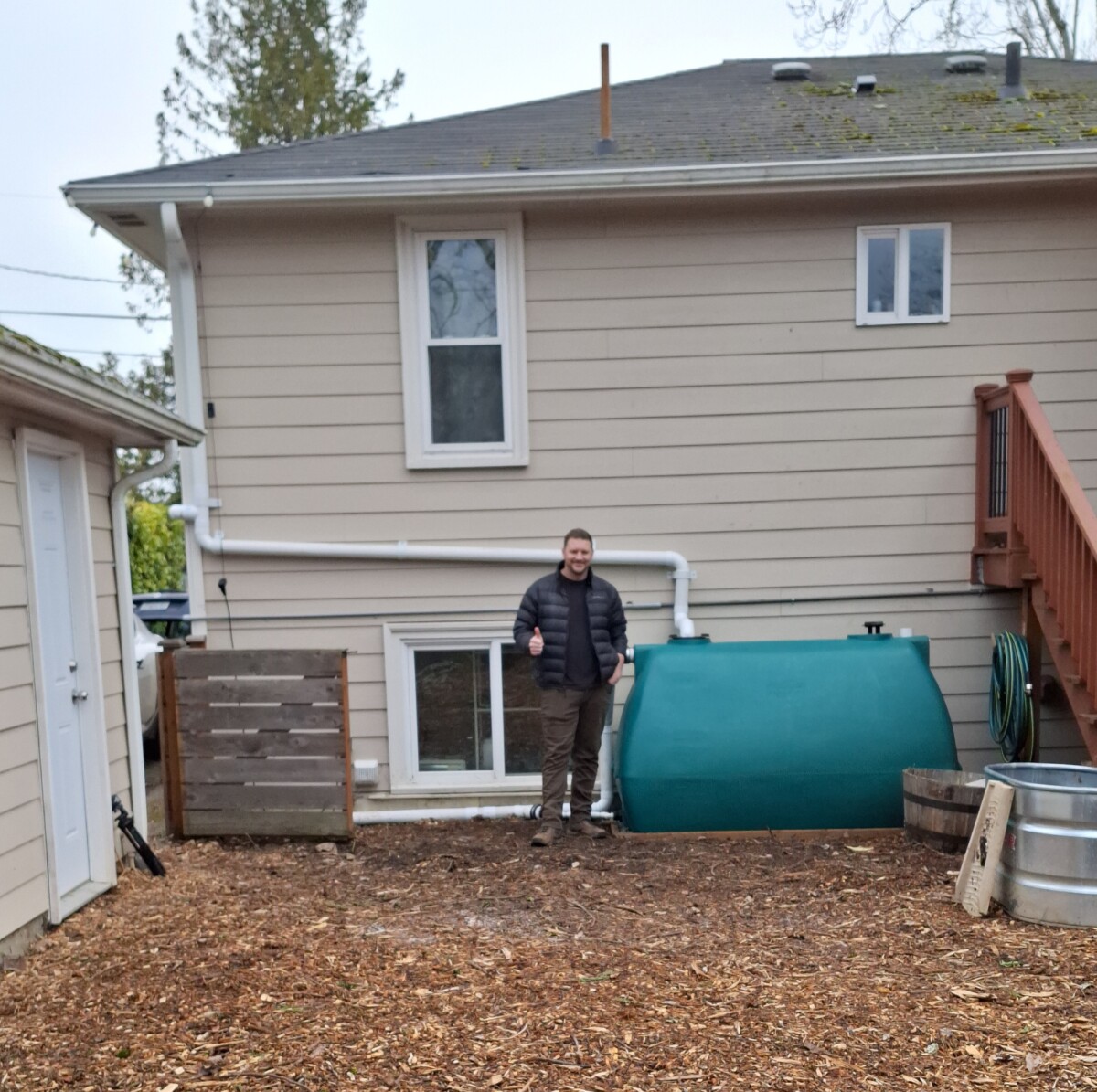 Homeowner standing beside a rain tank on a bed of bark in their backyard, highlighting sustainable landscaping practices